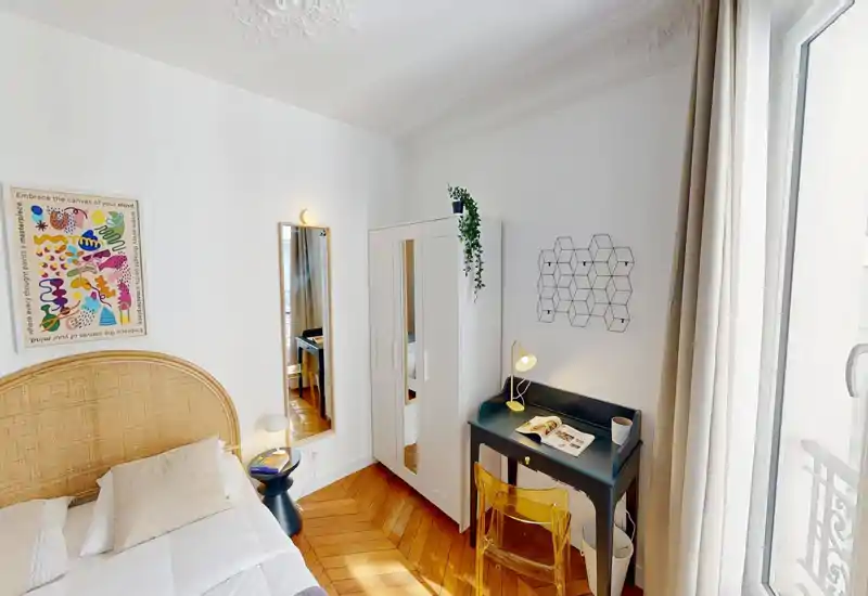 Chambre lumineuse et chaleureuse avec tête de lit en rotin, parquet et un petit bureau près de la fenêtre — idéale pour travailler ou se détendre.