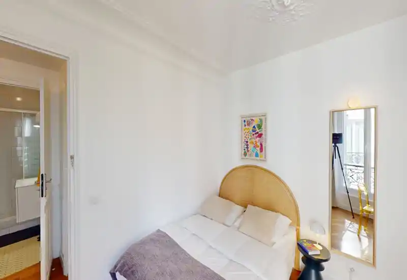 Chambre compacte et lumineuse avec une tête de lit en rotin, une literie blanche et une grande glace reflétant la lumière naturelle. L'espace est accueillant avec une table de chevet et des textiles doux.