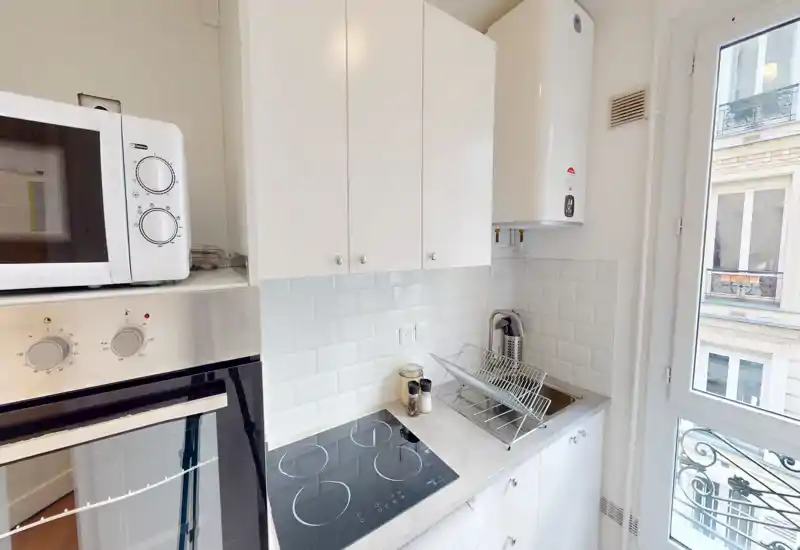 Kitchenette moderne et compacte avec four encastré, plaques vitrocéramiques, micro-ondes et petit évier; carreaux blancs et fenêtre pour une atmosphère lumineuse et soignée.