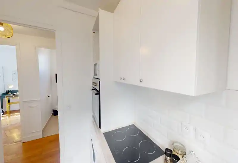 Cuisine compacte et moderne avec des placards blancs et une plaque de cuisson électrique, crédence carrelée lumineuse et petits accessoires sur le plan de travail ; le couloir adjacent et le sol en bois ajoutent de la chaleur.