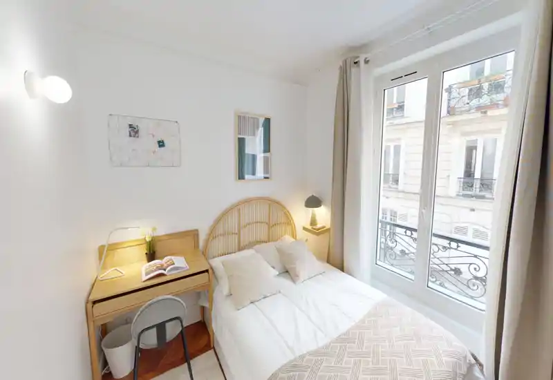 Chambre lumineuse et cosy avec tête de lit en rotin, lampe de chevet et bureau près de la fenêtre. La grande fenêtre apporte beaucoup de lumière et une vue agréable sur la rue.