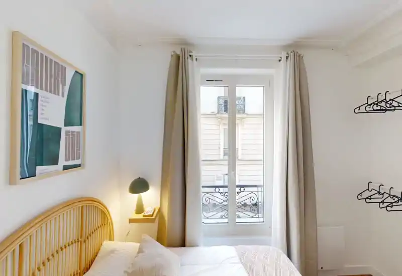 Chambre lumineuse et compacte avec tête de lit en rotin, lampe de chevet et grande fenêtre laissant entrer la lumière naturelle. Linge de lit neutre et décoration simple pour une ambiance calme et accueillante.