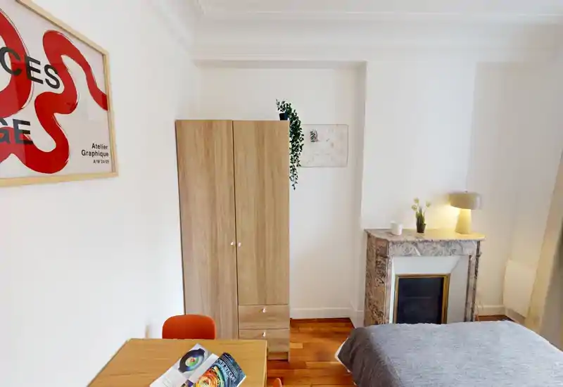 Chambre lumineuse et bien rangée avec lit double, armoire en bois et une cheminée décorative. L'atmosphère est accueillante grâce à une lampe de chevet et une décoration moderne épurée.