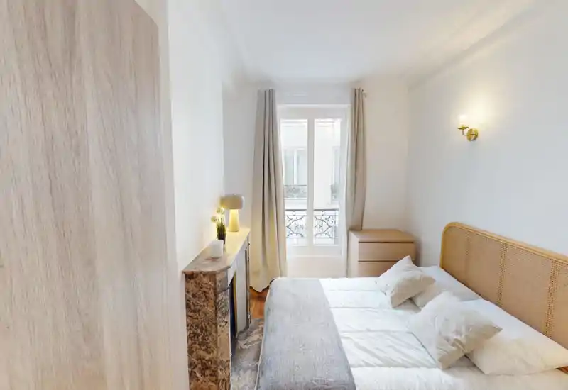 Chambre lumineuse et élégante avec un grand lit double, du linge de lit doux, une cheminée en marbre décorative, une lampe de chevet et la lumière naturelle d'une haute fenêtre avec rideaux.
