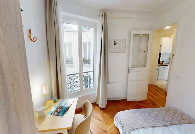 Chambre lumineuse et accueillante avec lit double, parquet, bureau et grande fenêtre laissant entrer la lumière naturelle. Décor épuré et moderne aux tons neutres avec une lampe chaleureuse.