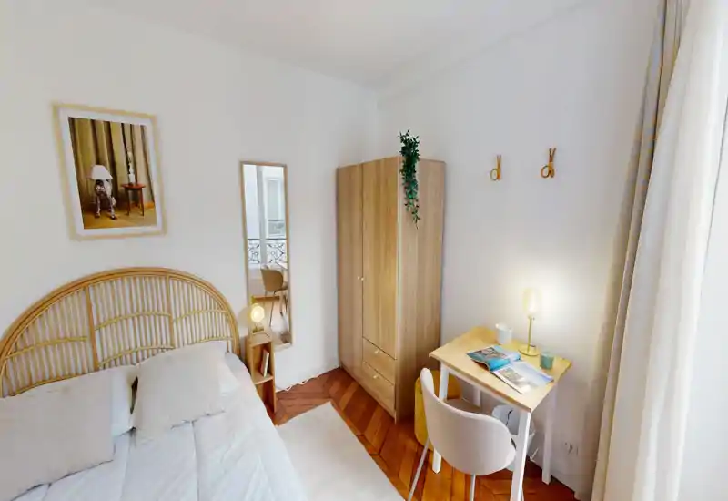 Chambre lumineuse et chaleureuse avec tête de lit en rotin, lampes de chevet et un petit bureau près de la fenêtre. Parquet clair et textiles neutres pour une ambiance accueillante, parfaite pour les séjours courts.