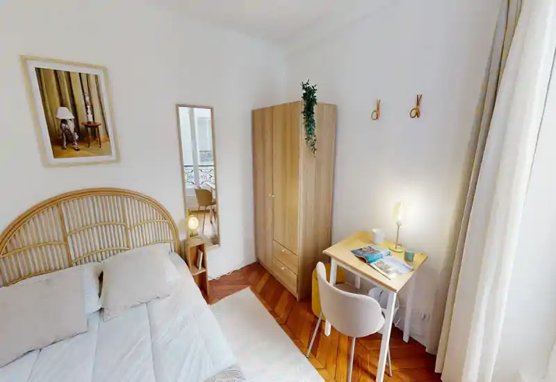 Chambre lumineuse et soignée avec tête de lit en rotin, literie bien faite, armoire et petit bureau près de la fenêtre — espace chaleureux et accueillant.