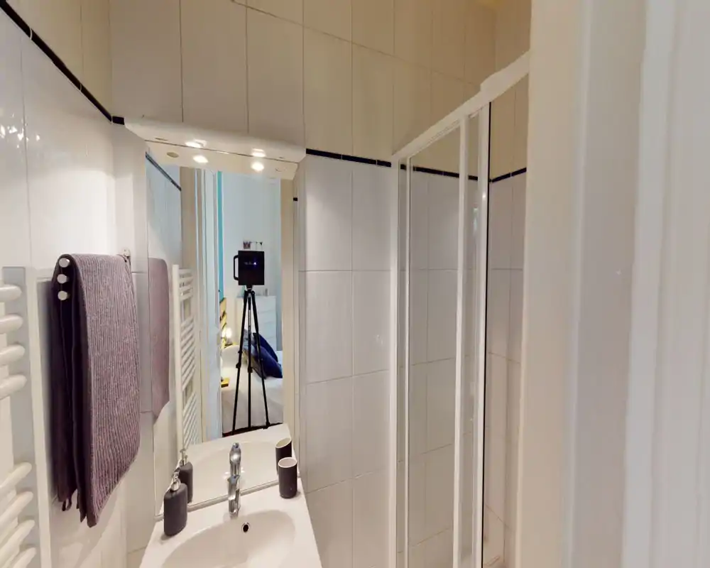 Baño alicatado y bien iluminado con lavabo, espejo y cabina de ducha en la esquina; toallas ordenadas y accesorios modernos.