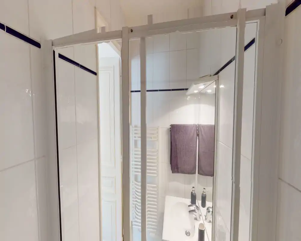Baño pequeño y bien iluminado con azulejos blancos, lavabo con grifería cromada, espejo y dos toallas moradas colgadas sobre el radiador.
