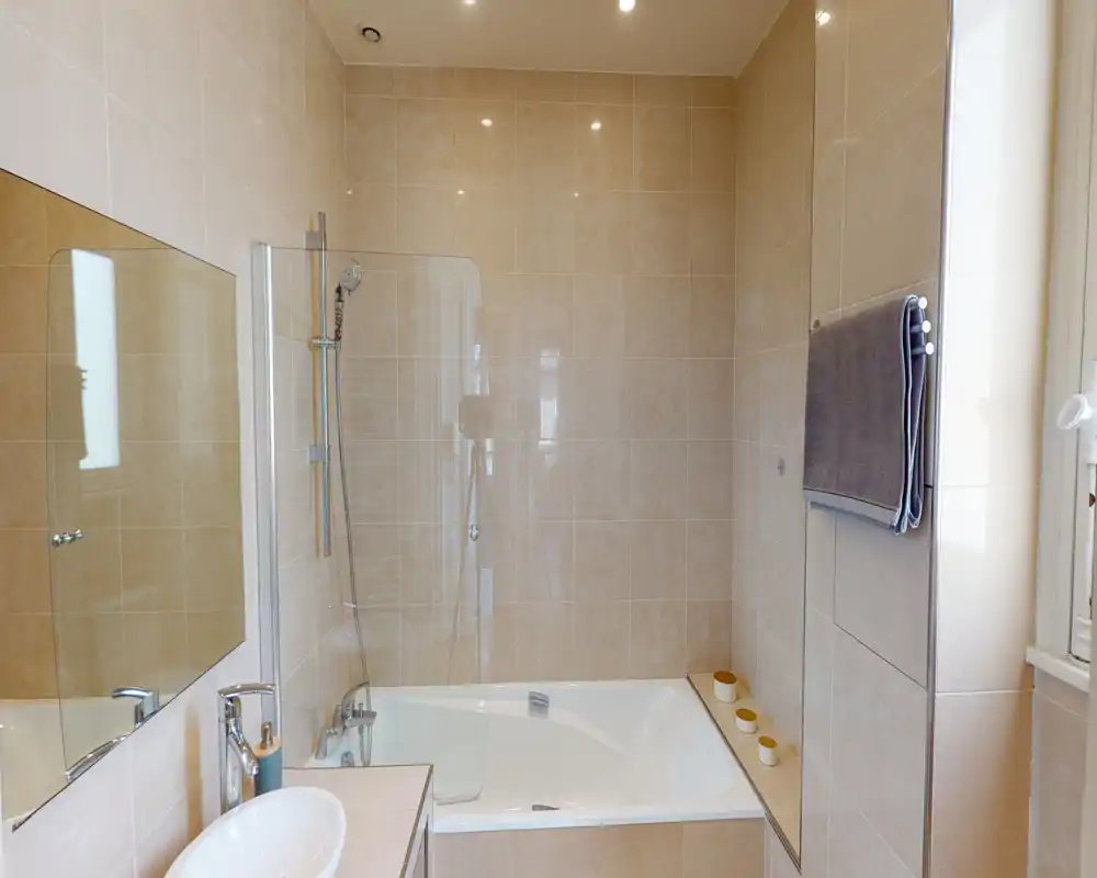 Salle de bains moderne carrelée avec baignoire et douche murale, pare-douche en verre, lavabo avec robinet chromé et porte-serviette — espace lumineux et propre.
