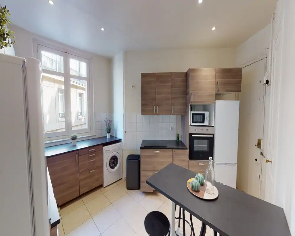 Cuisine moderne et lumineuse avec meubles imitation bois, four et micro-ondes intégrés, réfrigérateur grand format et un bar petit-déjeuner — grande fenêtre apportant une belle lumière naturelle.
