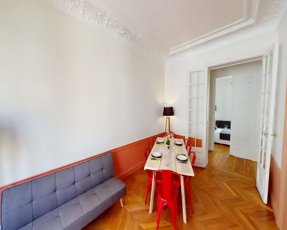 Coin repas lumineux avec une grande table en bois dressée pour six, chaises métalliques rouges et lampe sur pied décorative ; parquet élégant et moulures au plafond.