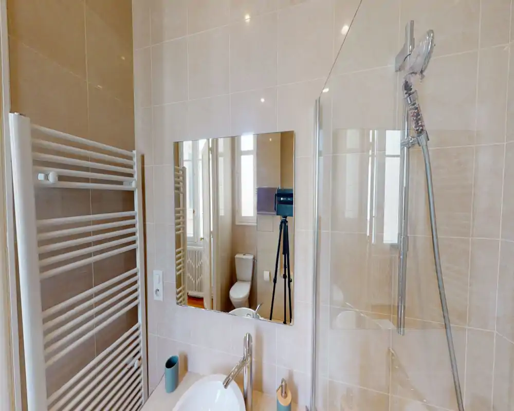 Salle de bains moderne et lumineuse avec murs carrelés, lavabo et robinet, paroi de douche en verre avec douchette et radiateur porte-serviettes mural. Le miroir reflète les toilettes et une fenêtre, agrandissant visuellement l’espace.