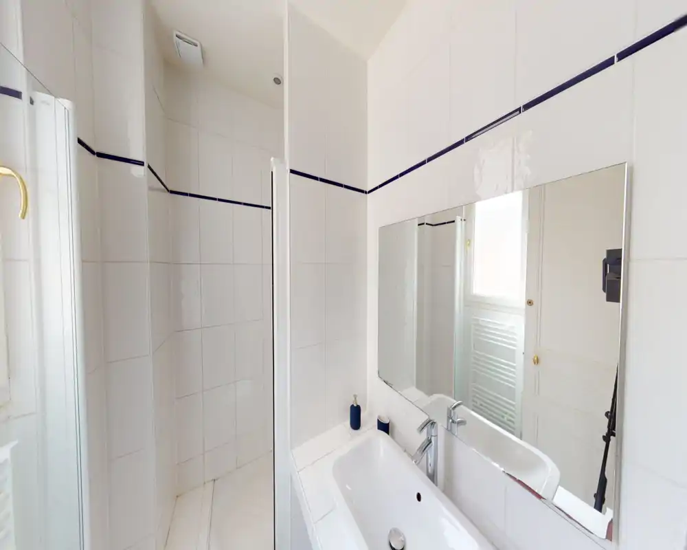 Salle de bains lumineuse tout en blanc avec douche à l'italienne, carrelage mural et lavabo avec grand miroir. Propre, minimaliste et bien éclairée, adaptée aux photos d'annonce.