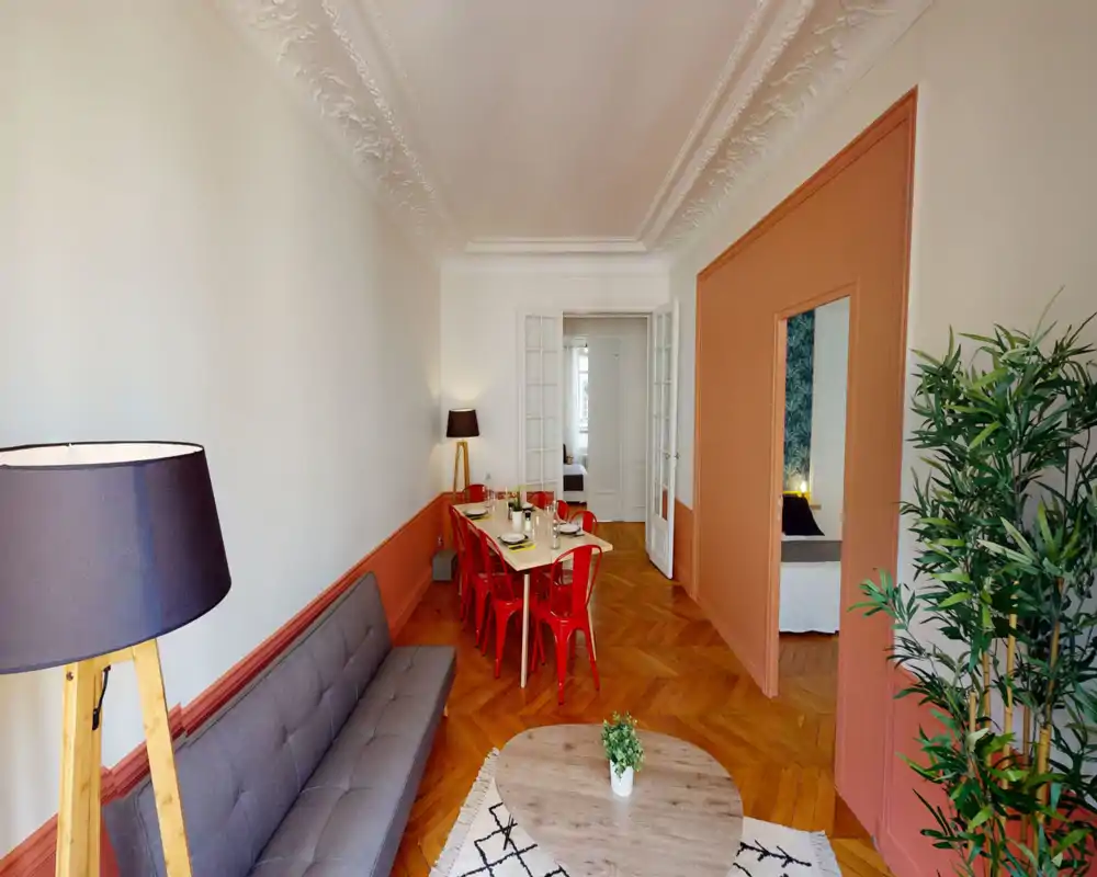 Espace séjour/salle à manger lumineux et élégant avec un canapé gris, table basse ronde et table à manger avec chaises rouges — parquet chaleureux et moulures décoratives raffinées.
