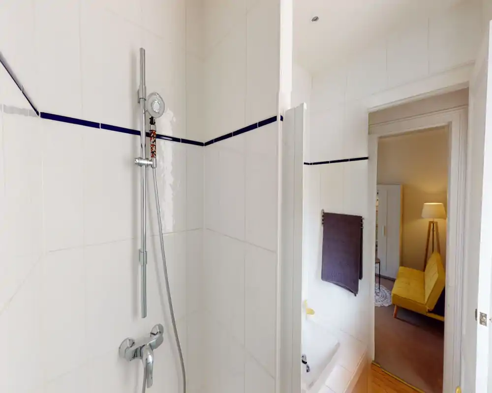 Salle de bains lumineuse et moderne avec douche carrelée et robinetterie chromée, baignoire partiellement visible ; serviette et porte donnant sur une pièce avec un canapé jaune.