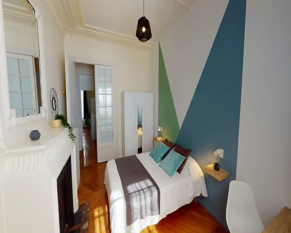 Camera da letto luminosa e di stile con letto matrimoniale completo, cuscini decorativi e una coperta accogliente. La stanza presenta pittura geometrica a parete, armadio, parquet e luce naturale soffusa.