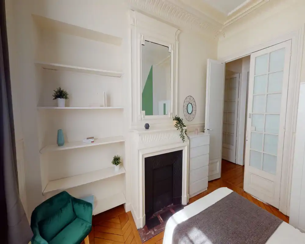Camera da letto luminosa ed elegante con camino decorativo, scaffali integrati e letto parzialmente visibile in primo piano. Pavimenti in legno, modanature raffinate e tocchi di verde che creano un'atmosfera accogliente e di stile, perfetta per annunci.