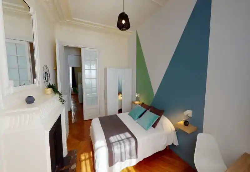 Camera da letto luminosa e di stile con letto matrimoniale completo, cuscini decorativi e una coperta accogliente. La stanza presenta pittura geometrica a parete, armadio, parquet e luce naturale soffusa.