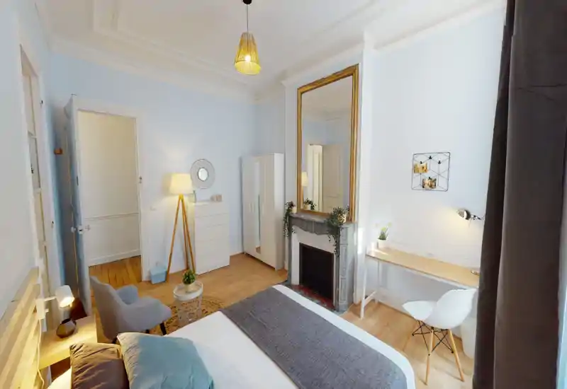 Camera da letto luminosa e di stile con un comodo letto matrimoniale, un angolo relax e una scrivania vicino alla finestra. Grande specchio sopra il camino decorativo e luce calda creano un’atmosfera accogliente.