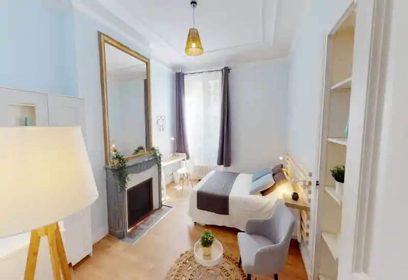 Camera da letto luminosa e di stile con letto matrimoniale, biancheria morbida, cuscini decorativi e un'area salotto accogliente. La luce naturale dalla finestra, lo specchio elegante e il pavimento in legno creano un'atmosfera invitante perfetta per gli annunci.