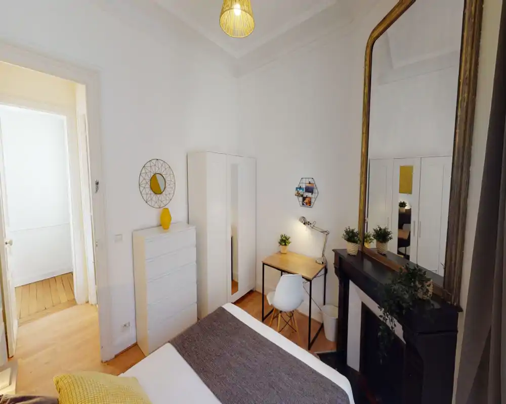 Bright, stylish bedroom with a large bed in the foreground, a white wardrobe and dresser, a small wooden desk with lamp, and a tall decorative mirror above a black mantel. Warm yellow accents and plants create a cozy, modern atmosphere ideal for rental photos.