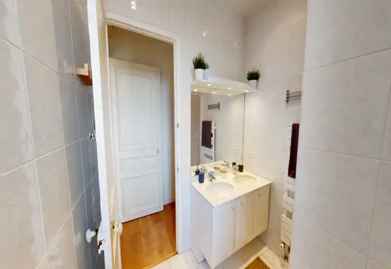 Salle de bains lumineuse et propre avec double vasque, grand miroir, radiateur porte-serviettes et carrelage mural ; agencement fonctionnel.