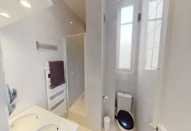 Salle de bains carrelée et compacte avec cabine de douche, WC à abattant sombre, plan vasque à double évier, sèche-serviettes et fenêtre dépoli apportant de la lumière naturelle.