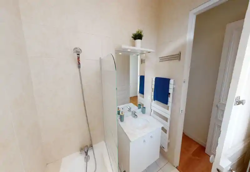 Petite salle de bain lumineuse avec baignoire et douchette, plan vasque effet marbre, miroir et radiateur porte-serviettes avec serviette bleue.
