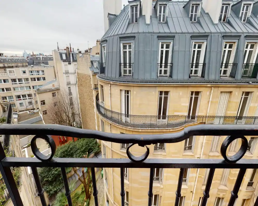 Vue depuis un petit balcon donnant sur un immeuble de style parisien avec toit mansardé et une cour en contrebas ; rambarde en fer forgé au premier plan.