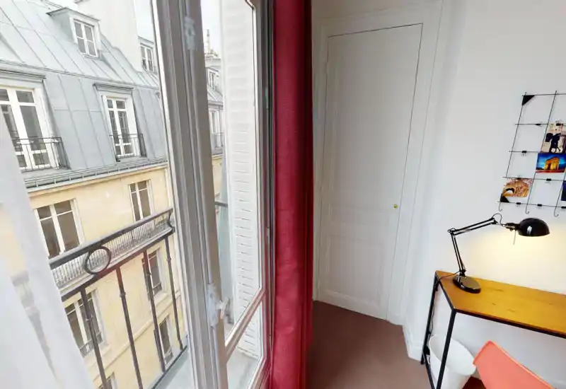 Coin lumineux d'un séjour avec une grande fenêtre donnant sur le balcon, un bureau simple avec lampe et une grille de photos décorative — idéal pour travailler ou lire.