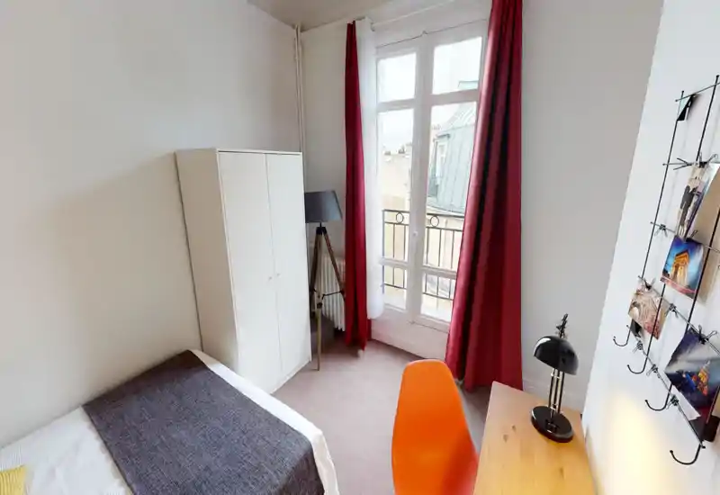 Chambre compacte et lumineuse avec un lit simple, une armoire, un bureau et une grande fenêtre avec rideaux rouges laissant entrer la lumière naturelle.