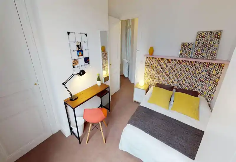 Chambre lumineuse et élégante avec un lit double encadré par une tête de lit colorée, deux coussins jaunes et un éclairage de chevet. Un petit bureau avec lampe et chaise offre un espace de travail; murs neutres et moquette douce pour un cadre chaleureux.