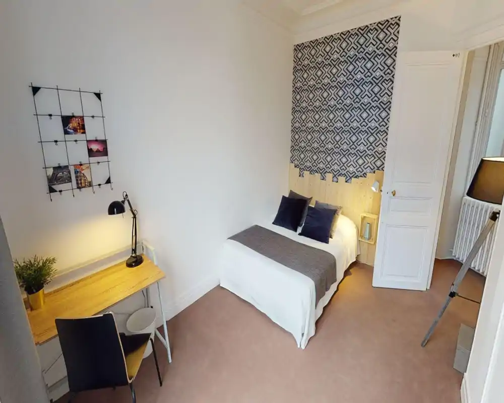 Chambre confortable et bien aménagée avec lit double, tête de lit décorative et mur à motif ; petit bureau et éclairage chaleureux, idéale pour invités ou étudiants.