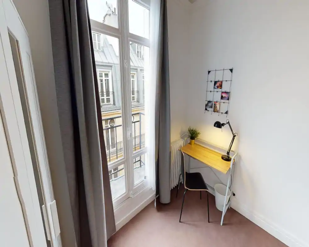 Coin lumineux et soigné servant de séjour/bureau avec un bureau près d'une grande fenêtre qui inonde la pièce de lumière ; décoration simple avec lampe et plante pour une touche chaleureuse.