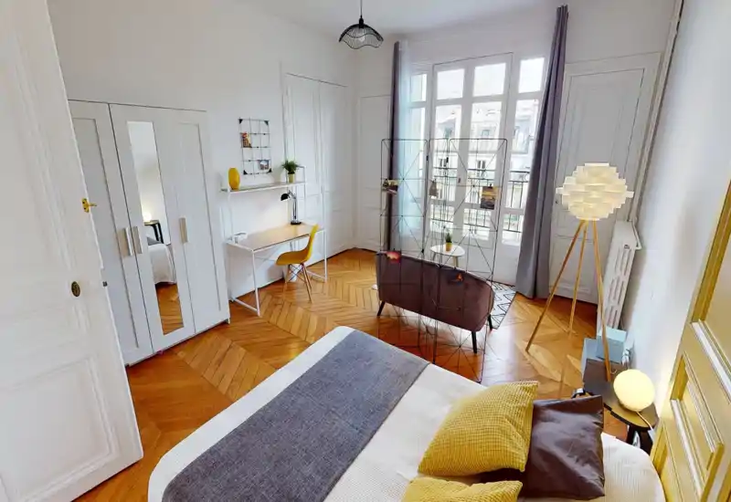 Bright, stylish bedroom with a large bed in the foreground, warm herringbone wood floors, a wardrobe and a small seating area by tall windows letting in abundant natural light.