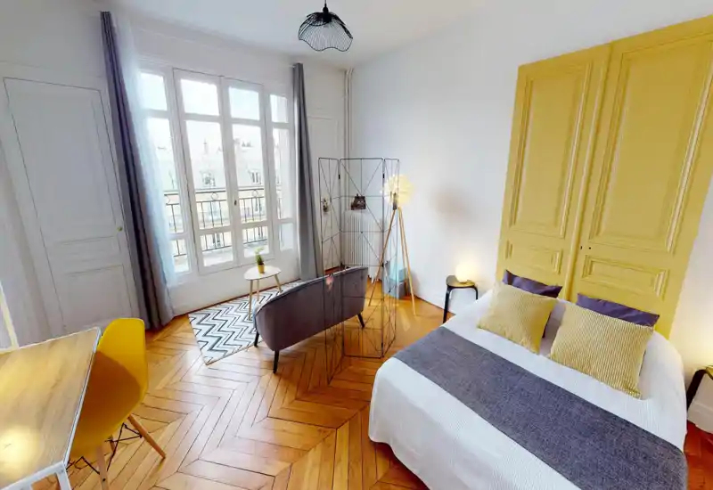 Bright, stylish bedroom with a comfortable double bed, decorative yellow panel headboard, wooden herringbone floor and large windows providing plenty of natural light. A small seating area with a sofa and coffee table creates a cozy corner.