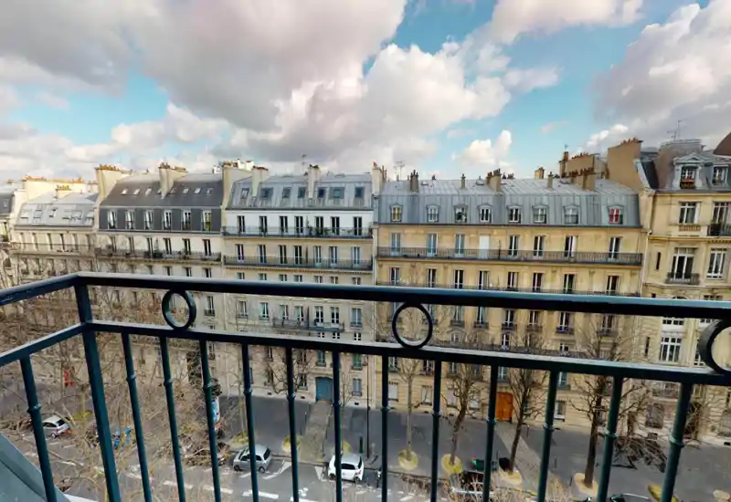 Balcony view overlooking a Parisian street and classic Haussmann-style buildings, with a metal railing in the foreground and a bright sky above.