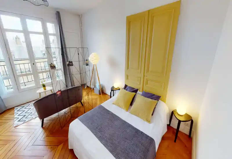 Bright, stylish bedroom with a full bed in the foreground, decorative pillows and layered textiles; large windows provide abundant natural light and a small seating area is visible behind a decorative room divider.