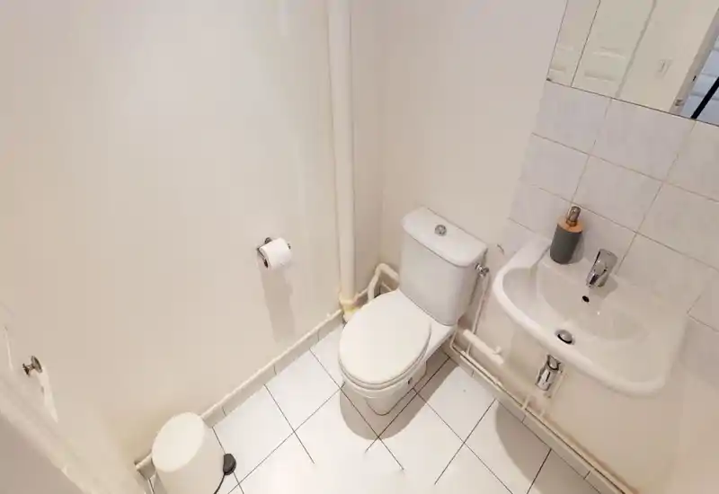 Petite salle de bain propre avec WC, lavabo mural, crédence carrelée et sol carrelé blanc — bien éclairée et rangée.