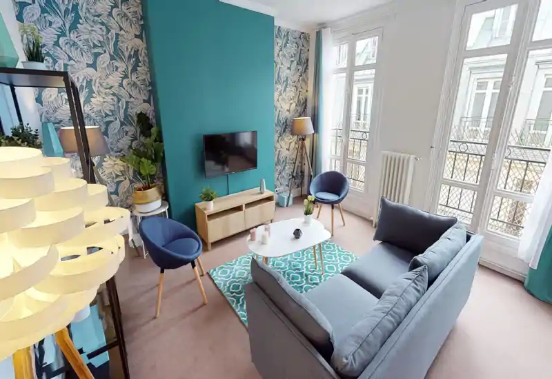 Séjour lumineux et élégant avec un canapé gris confortable, deux fauteuils bleus, un tapis à motifs et de grandes fenêtres apportant beaucoup de lumière naturelle — idéal pour se détendre ou recevoir.