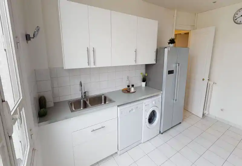 Cuisine lumineuse et propre avec placards blancs, double évier, lave-vaisselle et lave-linge encastrés, et grand réfrigérateur inox. Quelques éléments décoratifs apportent une ambiance moderne et fraîche, parfaite pour une annonce.