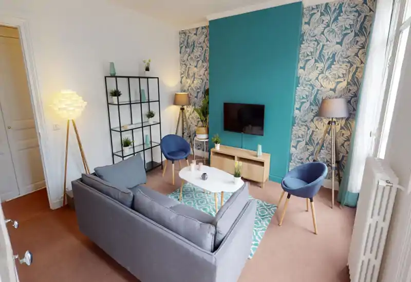 Salon lumineux et élégant avec canapé gris, deux fauteuils bleus modernes, tapis à motifs et télévision murale. Lampadaires chaleureux et accents turquoise soignés créent une ambiance cosy et contemporaine.