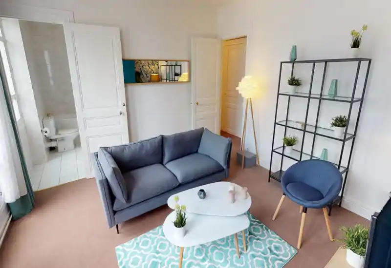 Salon lumineux et stylé avec canapé deux places, fauteuil d'appoint, tables basses et étagère décorative — bien éclairé par la lumière naturelle et une lampe sur pied.