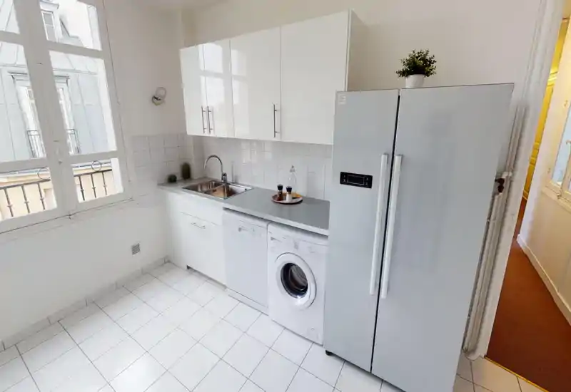 Cuisine moderne et lumineuse avec meubles blancs, évier double, lave-vaisselle et lave-linge, ainsi qu’un grand réfrigérateur. Sol carrelé propre et grande fenêtre pour une belle lumière naturelle.