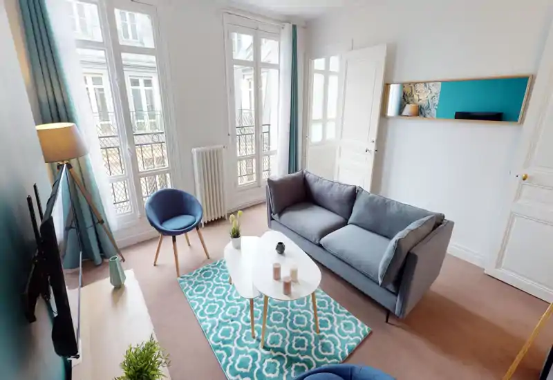 Salon lumineux et élégant avec un canapé gris confortable, tapis turquoise à motifs et lumière naturelle provenant de grandes fenêtres — un espace moderne et chaleureux.