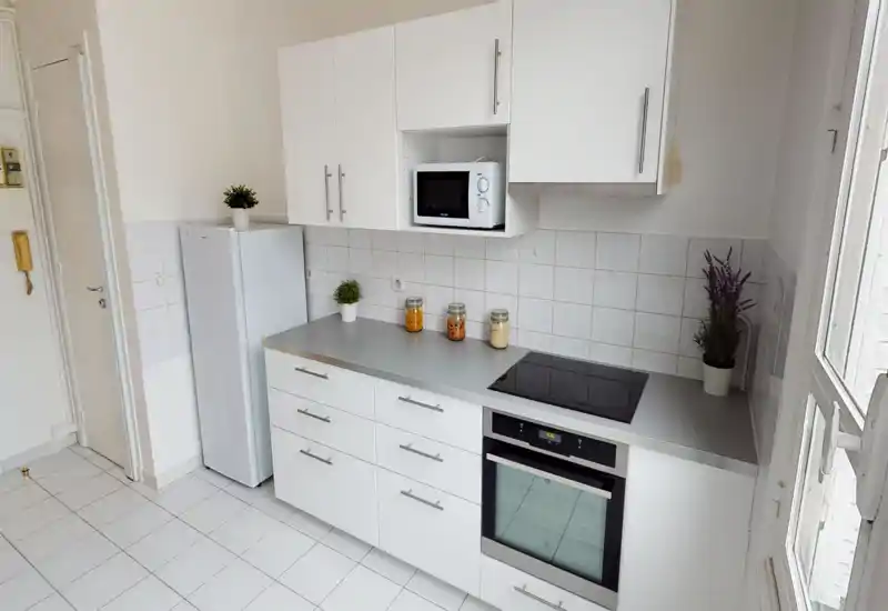 Cuisine moderne et lumineuse avec placards blancs, four encastré et plaque à induction, réfrigérateur et micro-ondes compact. Crédence carrelée propre, bocaux décoratifs et petites plantes pour une ambiance fraîche et soignée.