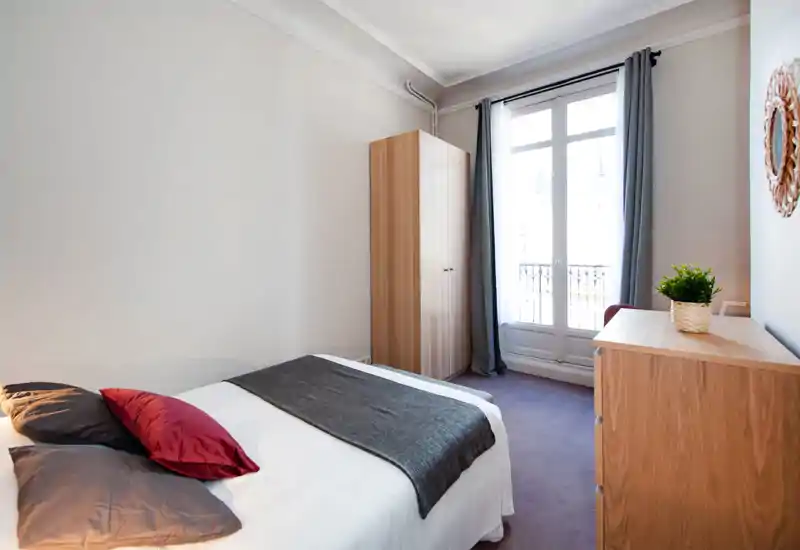 Bright, neatly staged bedroom with a double bed, decorative pillows, a wardrobe and a wooden dresser by a large window that admits generous natural light.