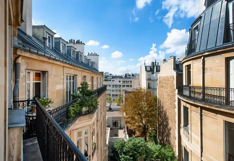 Sunny city balcony view overlooking classic Parisian-style buildings, leafy trees and a bright blue sky — great for listings highlighting an outdoor terrace or balcony.