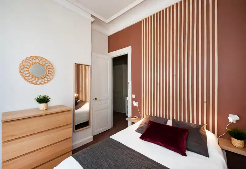 Bright, styled bedroom with a wooden slatted feature wall, comfortable double bed dressed with pillows and a velvet cushion, and minimalist wooden furniture including a chest of drawers and bedside shelves.